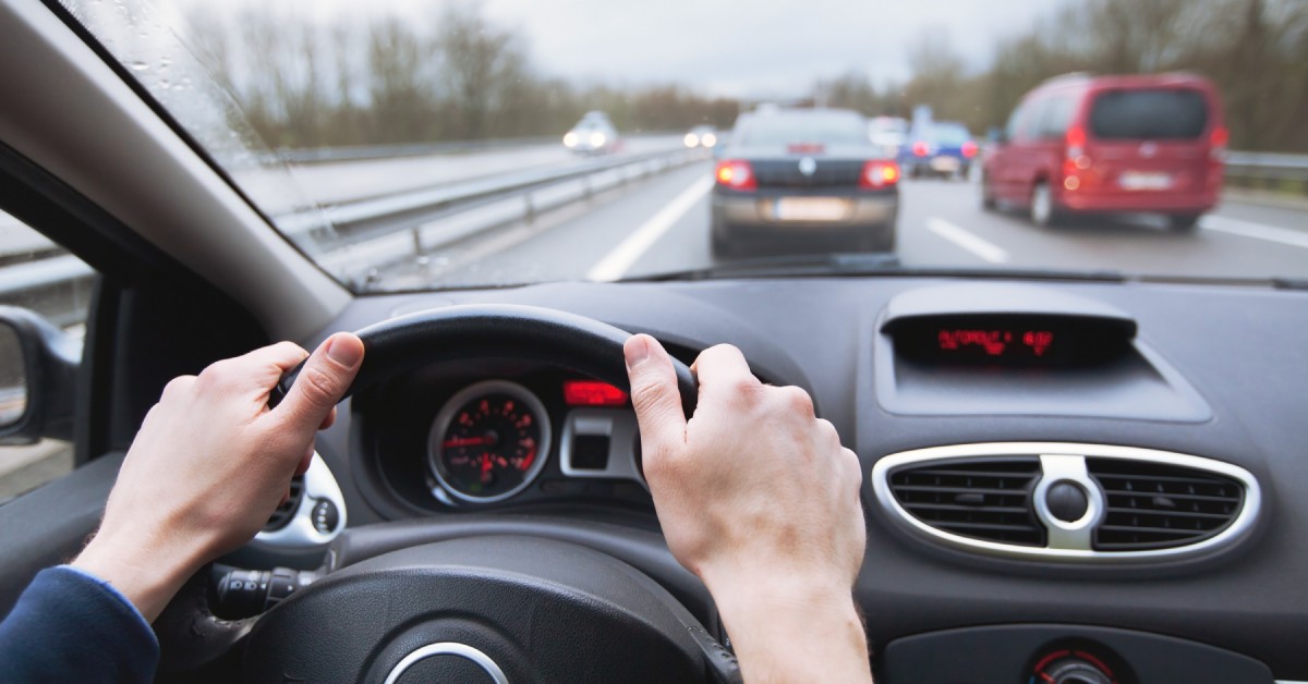Close-up of someone’s hands at the ten and two positions on a car’s steering wheel while driving on the highway.