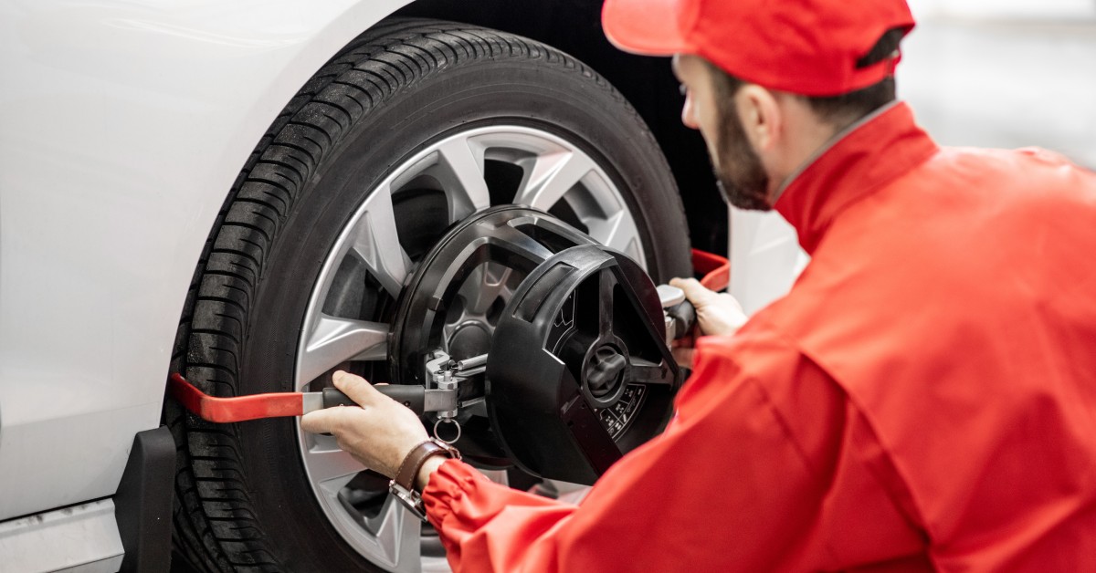An automotive technician in a red hat and coveralls uses a fixing disk for wheel alignment on a white car.