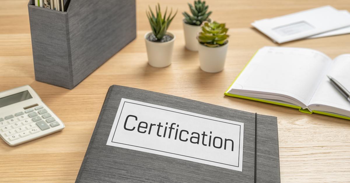 Close-up of a desk that has plants, notebooks, a calculator, and a folder on it that says “certification” on the front.