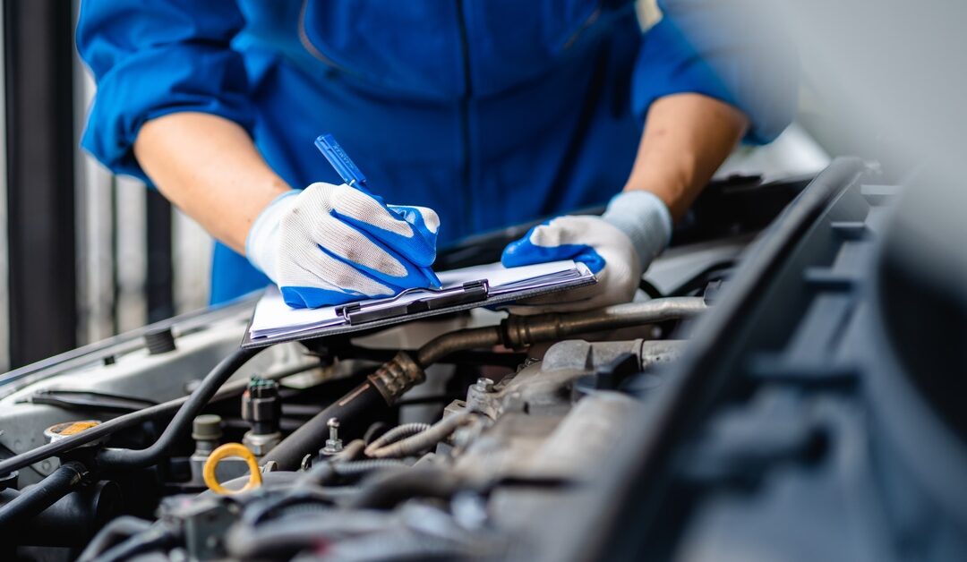 An automotive mechanic stands next to a car with the hood popped up. They are writing on a clipboard.