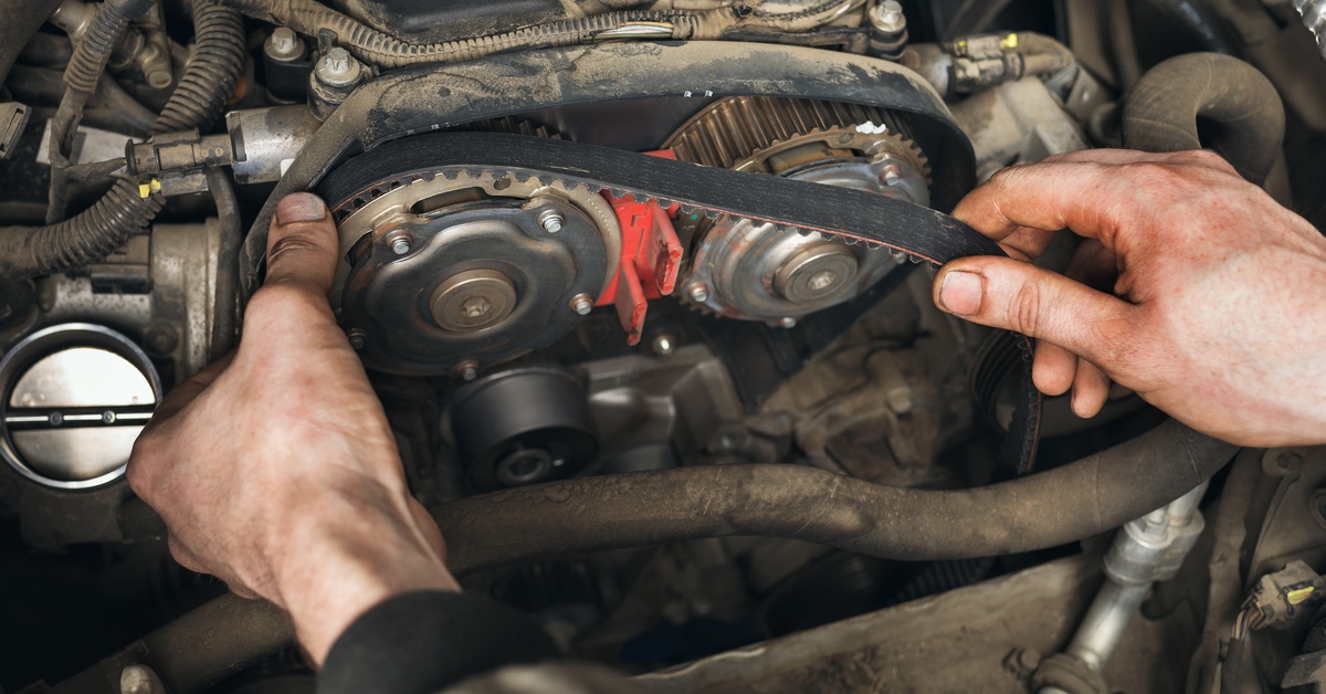 A mechanic with both hands under the hood of a car replacing a timing belt. They're setting it on the housing.