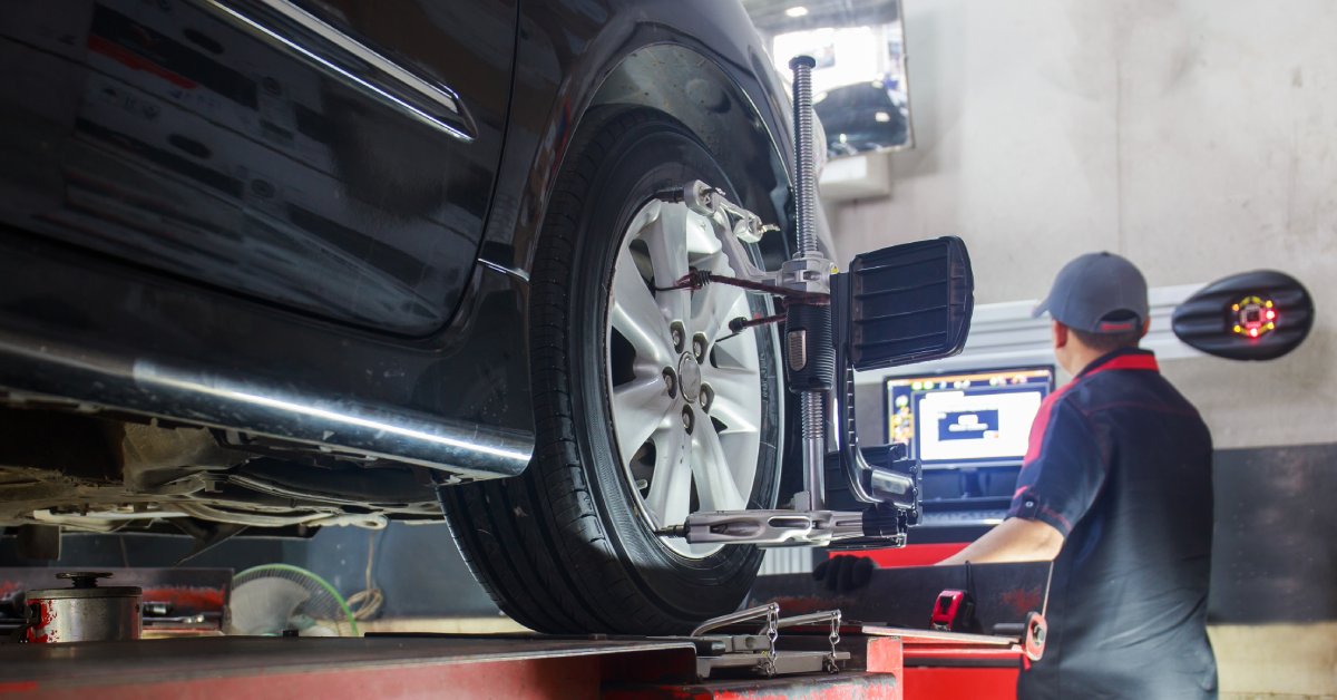 A mechanic stands at the computer to monitor a car's alignment process. The car is black with equipment attached to the tire.