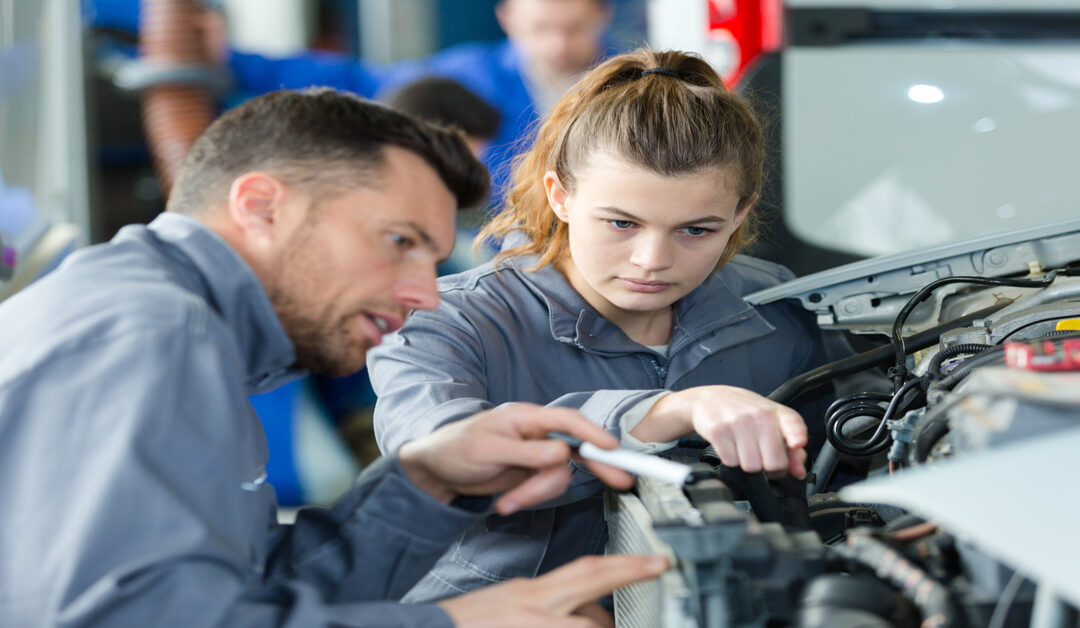 An older male mechanic looking at a car's components as he speaks to a young female student about the part.