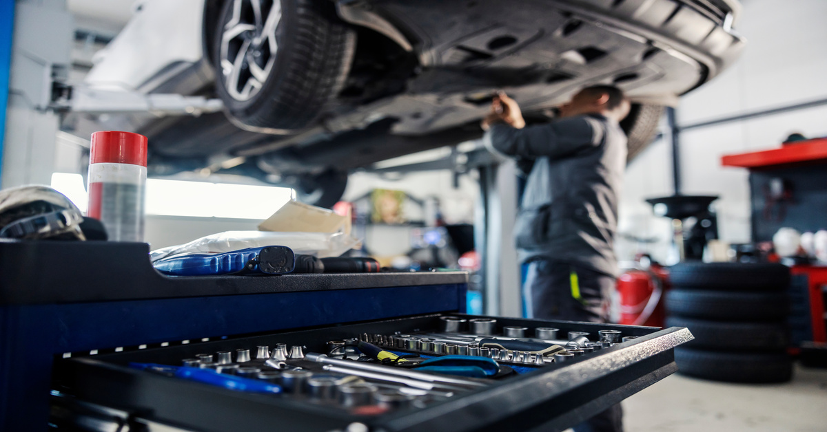 An automotive mechanic stands under a car as the car sits on a lift. There is a tool cart with a drawer open.