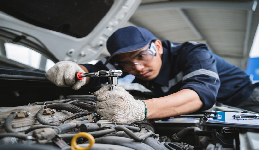 An auto mechanic in uniform with a baseball cap and gloves on. He is under the hood of a vehicle with wrenches.