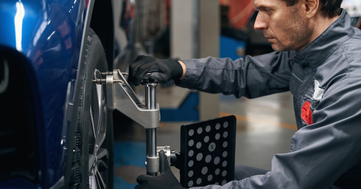 A man automotive technician works on a tire with a wheel alignment machine attached to the rim. He wears black gloves.

