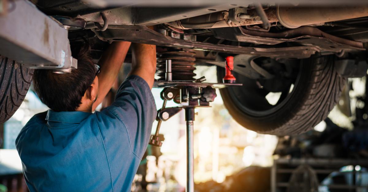 A mechanic under the body of a car that's been lifted. They have both arms inside the car's underbody for a repair.