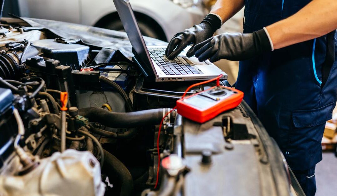 An automotive mechanic stands at the front end of a car with the hood open. They're researching on a laptop.