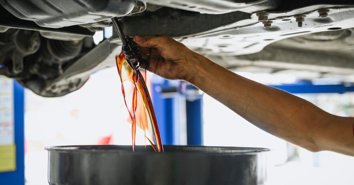 A mechanic's arm under the body of a car twisting the oil cap as the oil drains into a pan. The oil is brown in color.