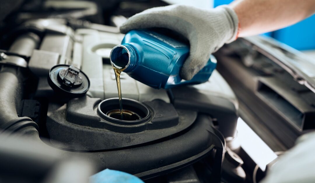 A mechanic under the hood of a car wears gloves as they pour a blue bottle of fluid into a reservoir.