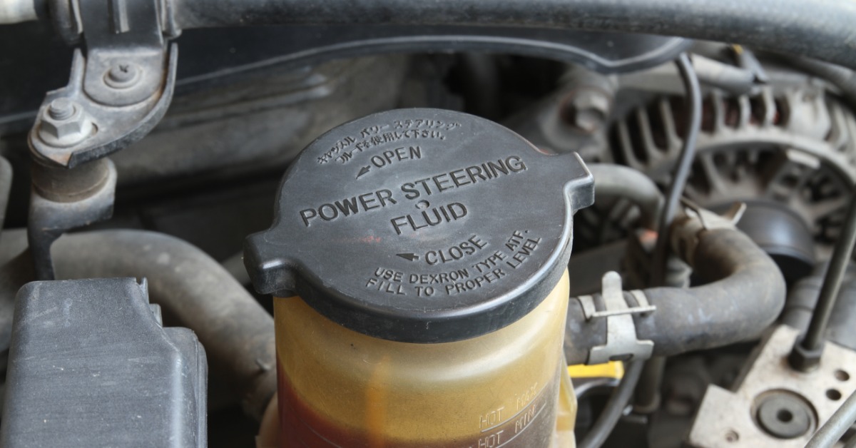 A close-up of a vehicle's power steering reservoir with the cap on. The fluid is brown and the cap is labeled.