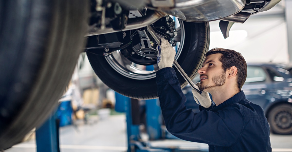 An automotive mechanic underneath a vehicle on a car lift. The mechanic wears a navy blue uniform and white gloves.